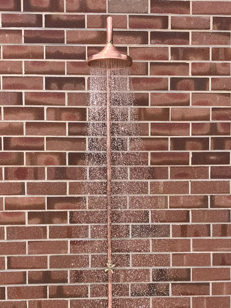 Copper outdoor shower against a classic red brick wall beside a green-framed window