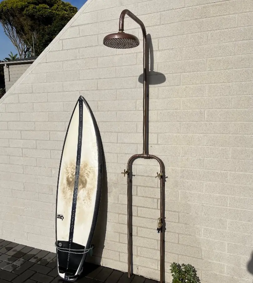 A surfboard leaning beside a full copper outdoor shower against a white brick wall